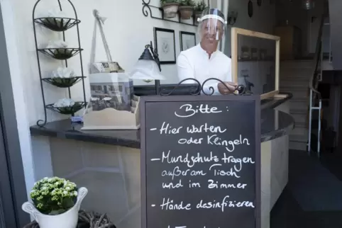 Oliver Jakob mit Schutzvisier an dem Schild, das auflistet, was alles beim Restaurantbesuch zu beachten ist.