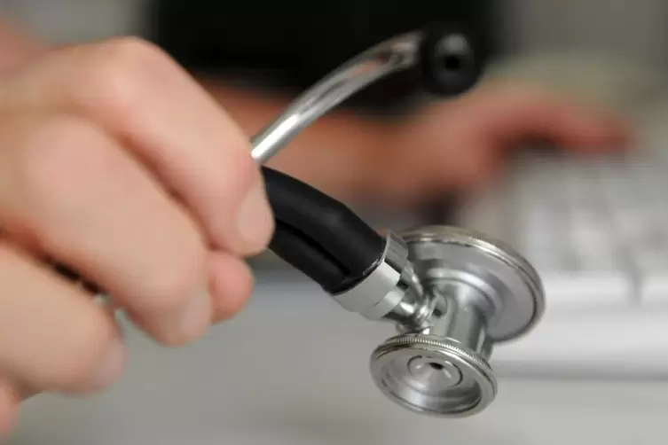 Ein Arzt hält in einem Behandlungszimmer in seiner Praxis ein Stethoskop in der Hand, mit der anderen Hand bedient er eine Compu