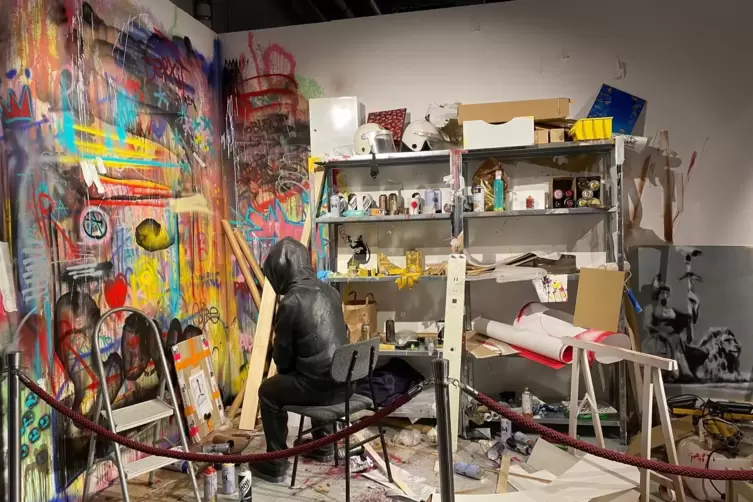 Banksy ist in seiner Ausstellung – sogar in seiner Werkstatt. Aber würde er da verhüllt und mit Kapuze sitzen? Seltsame Installa