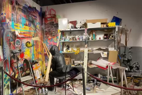Banksy ist in seiner Ausstellung – sogar in seiner Werkstatt. Aber würde er da verhüllt und mit Kapuze sitzen? Seltsame Installa