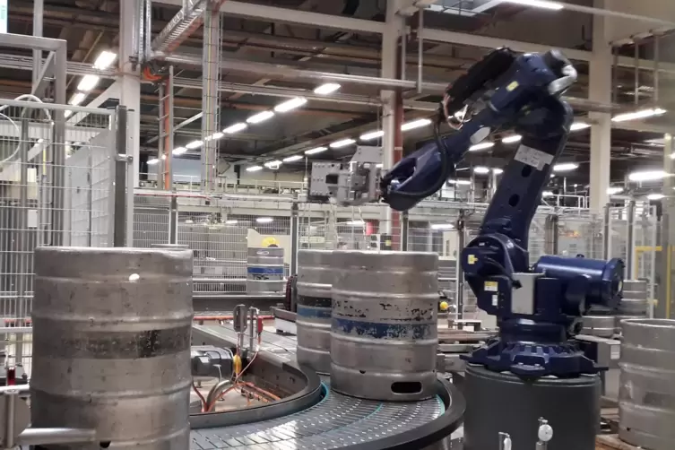 Ein blauer Roboterarm hebt die noch leeren Bierfässer zur Reinigung und zum Neu-Befüllen auf ein Förderband.