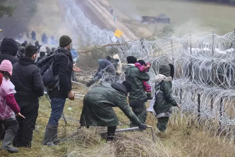 Migranten stoßen an der belarussich-polnischen Grenze einen Zaun um, während die Polizei hinter einem Stacheldrahtzaun steht.