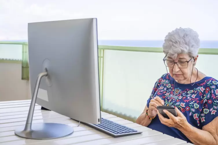 Keine Scheu vor Informationstechnologie ( IT): Eine Seniorin sitzt mit dem Smartphone in der Hand vor einem PC.