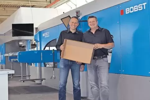 Geschäftsführer Carsten Zier (links) und Betriebsleiter Mathias Heiler vor der neuen Maschine, mit der bedruckte Stanzverpackung